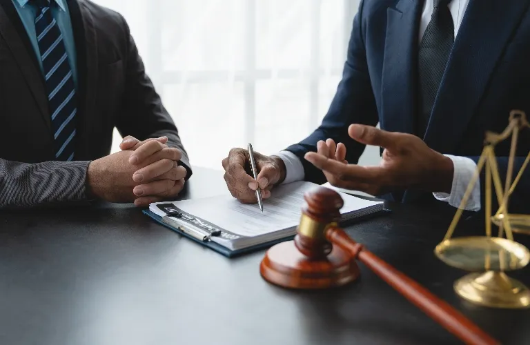 Two men in suits engaged in discussion over legal documents with gavel and scales of justice on table.