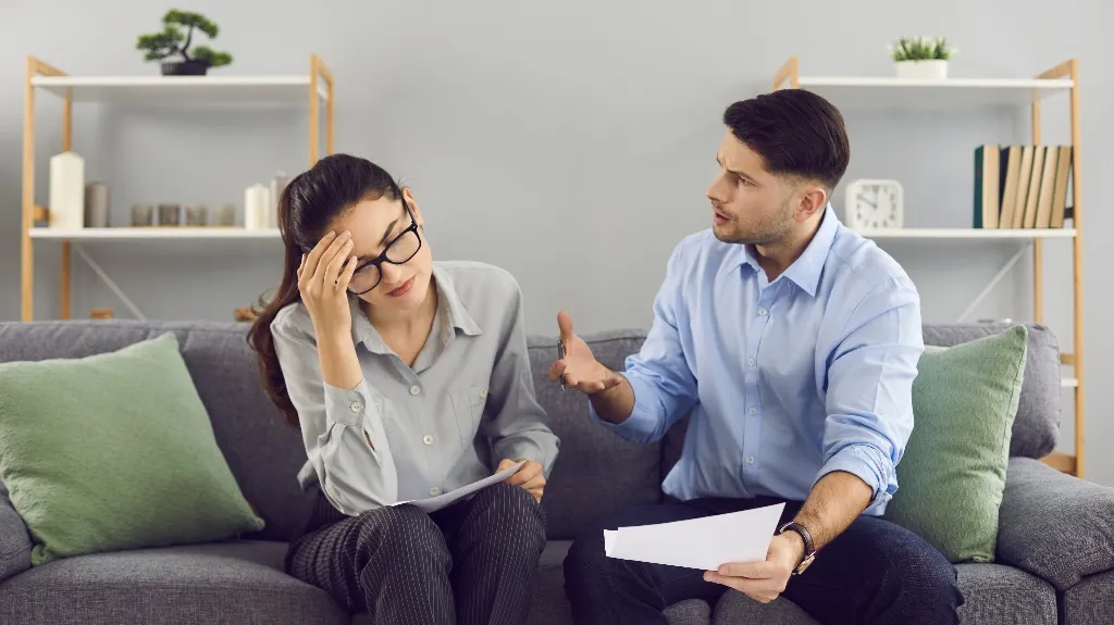 Two adults in casual attire sitting on a gray couch, discussing documents, appearing stressed.