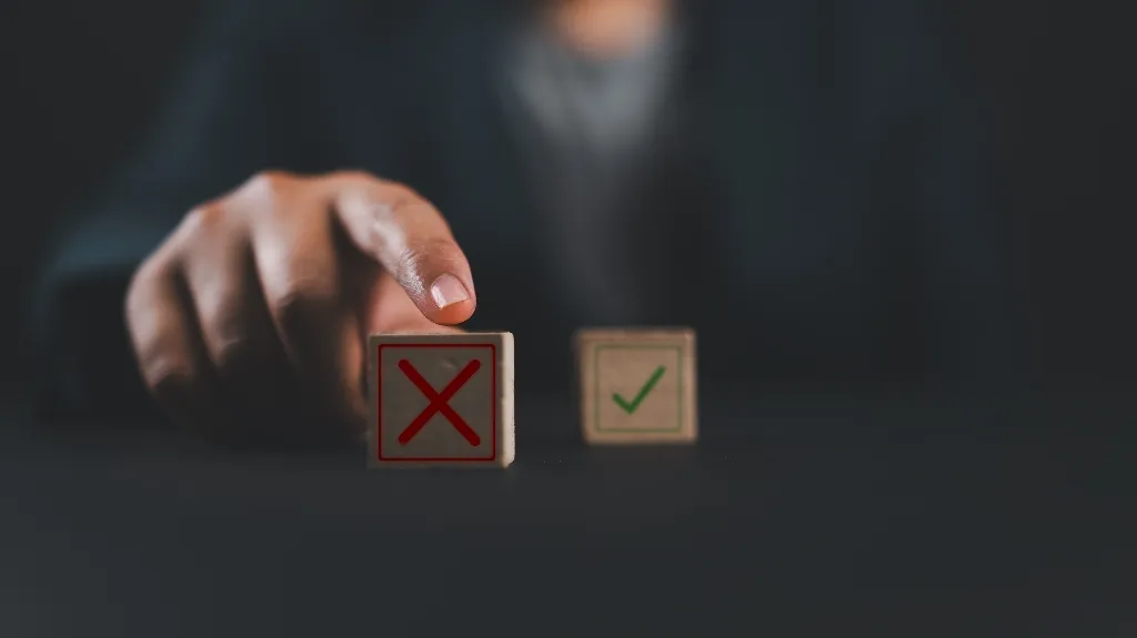 Hand pointing at wooden block with red X, green checkmark block in background on dark surface.