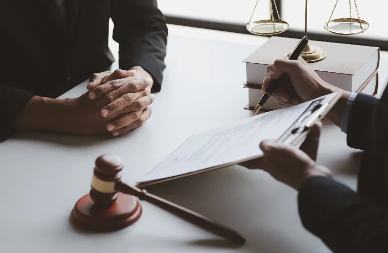 Two professionals in dark suits at a desk with legal books, a gavel, and a document on a clipboard.