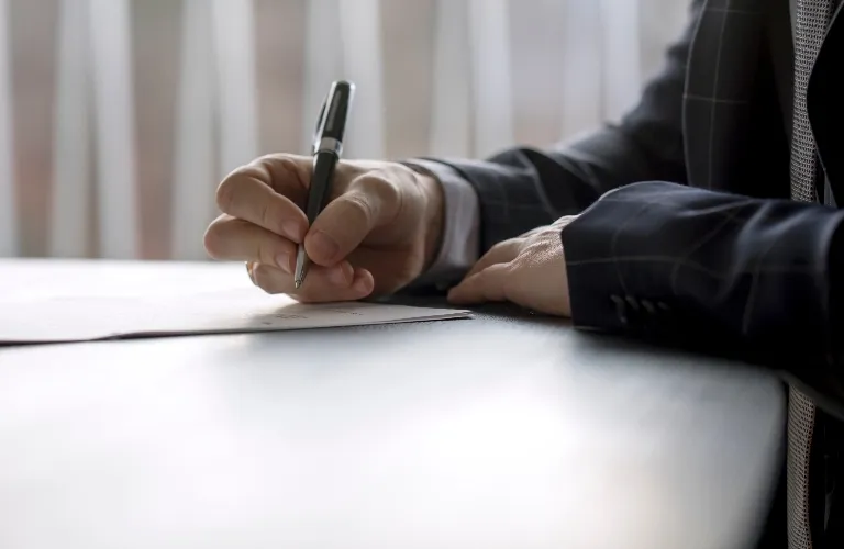 Person in dark suit writing with black pen on white paper at desk
