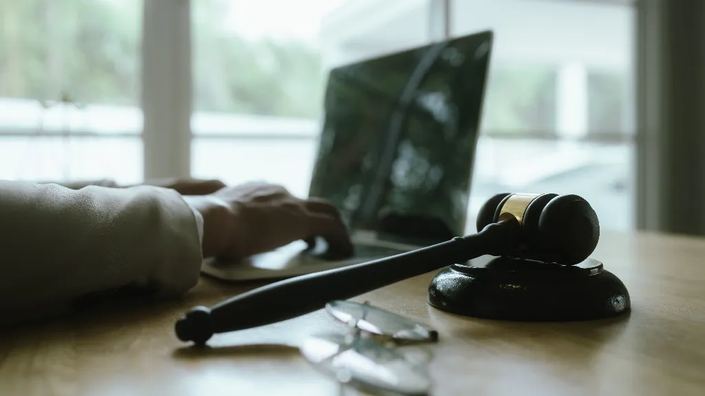 Close-up of a wooden judge's gavel on a desk with a person typing on a laptop in the background.