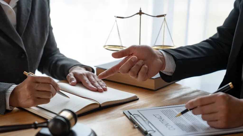 Two professionals in suits discussing and signing legal documents with scales of justice and gavel on desk.