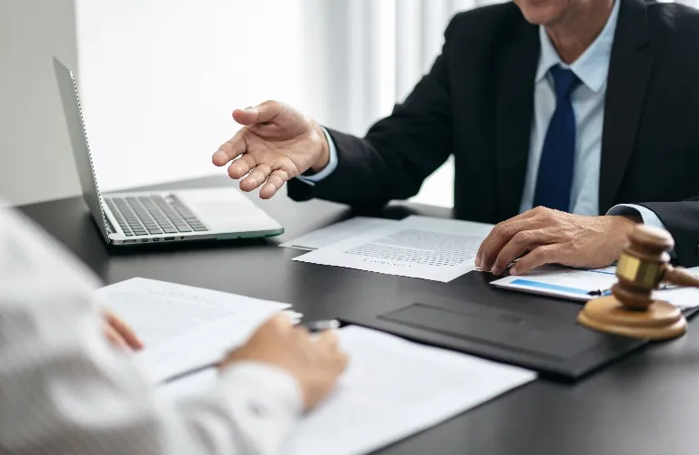 Two professionals in formal attire discussing documents at a desk with a laptop and a judge's gavel.