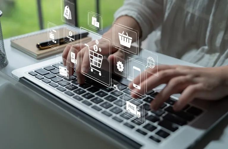 Person typing on laptop keyboard with transparent digital shopping and e-commerce icons.