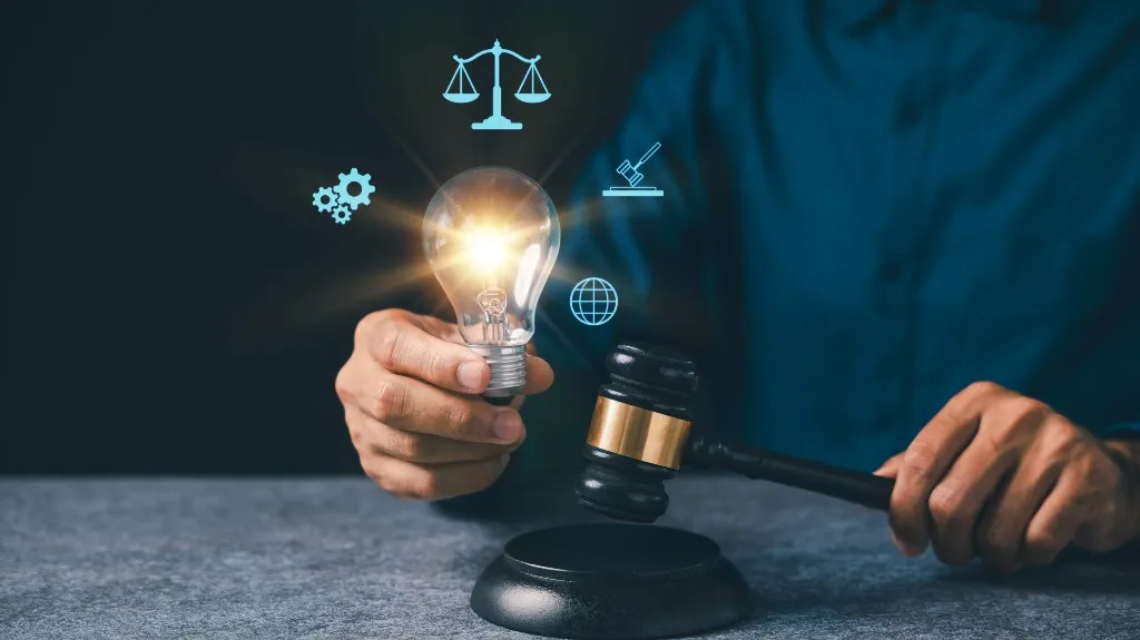 Person holding a glowing light bulb and a judge's gavel with legal icons surrounding the bulb.