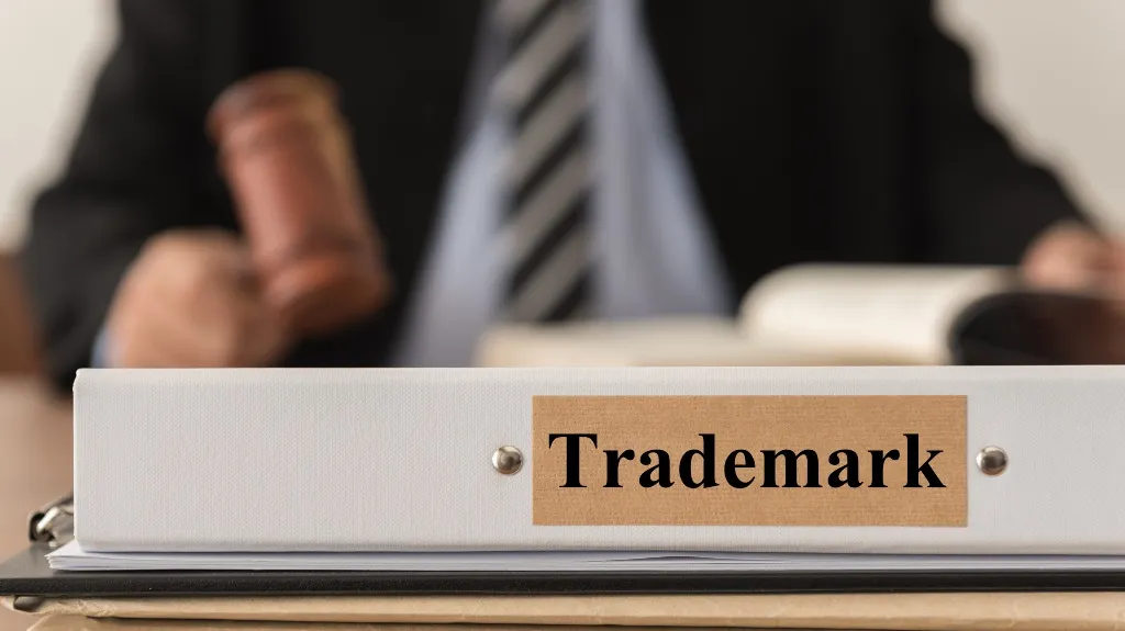Close-up of a white binder labeled "Trademark" with a blurred person holding a gavel in the background.